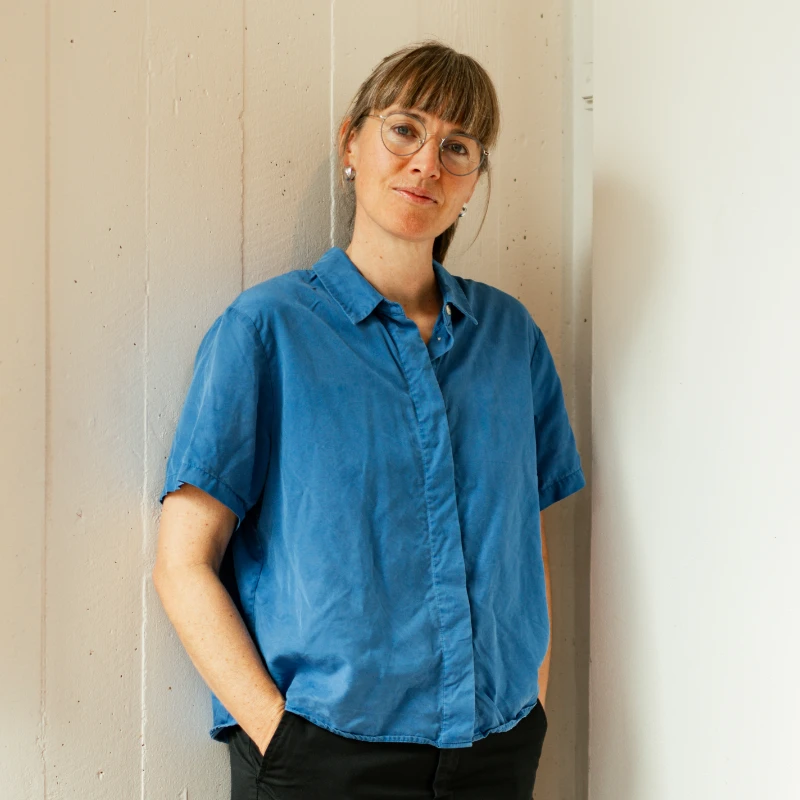 Mette Øyås Madsen i blå skjorte foran en lys væg.